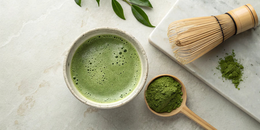 A ceramic bowl of matcha with a bamboo whisk and a spoon of matcha powder.